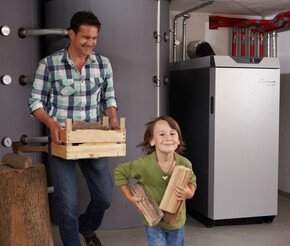Kind rennt lächelnd mit Holzstücken vom Vater weg, der eine Kiste mit Holz für die Holzheizung trägt | © bösch heizung.klima.lüftung