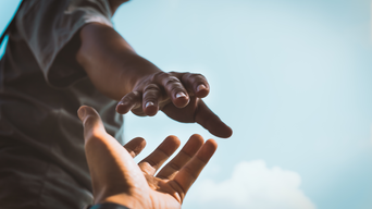 Dynamische Aufnahme aus der Untersicht zweier Hände, die sich gegenseitig zu greifen. Eine Hand streckt sich von unten nach oben, während die andere sich helfend entgegenstreckt, symbolisch für Unterstützung und Zusammenhalt, vor einem klaren blauen Himmel