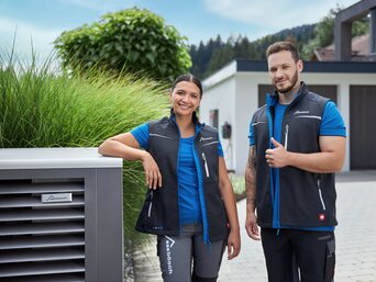 Zwei lächelnde Techniker, eine Frau und ein Mann, stehen neben einer Außeneinheit einer Wärmepumpe. Beide tragen Firmenkleidung mit dem Logo von bösch. Im Hintergrund ist ein modernes Wohnhaus sichtbar. Der Techniker hält einen Daumen hoch. | © bösch heizung.klima.lüftung