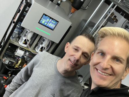 Besitzer Stefan V. und ein bösch Techniker lächeln in die Kamera vor einem komplexen Heizungssystem in einem technischen Raum. | © Walter Bösch GmbH & Co KG