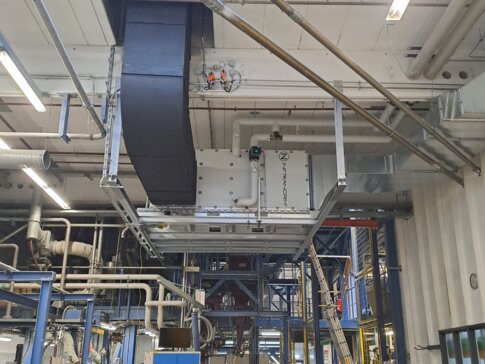 Industrielle Produktionshalle mit komplexen Rohrleitungen und einer großen, schwarzen Lüftungsanlage an der Decke. Unten arbeitet eine Person an einem Tisch mit einem Laptop, umgeben von Maschinen und technischen Geräten. Im Hintergrund sind Leitern und weitere technische Strukturen zu sehen. | © Infranorm, Wels (AT)