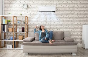 Frau sitzt mit Fernbedienung auf Couch, aus der Klimaanlage kommt frische Luft