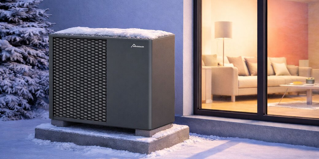 Monoblock-Luft-Wasser-Wärmepumpe von bösch im Winter, mit Schnee bedeckt, auf Betonsockel neben Fensterfront eines modernen Einfamilienhauses