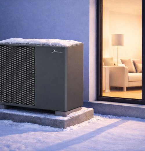 Monoblock-Luft-Wasser-Wärmepumpe von bösch im Winter, mit Schnee bedeckt, auf Betonsockel neben Fensterfront eines modernen Einfamilienhauses