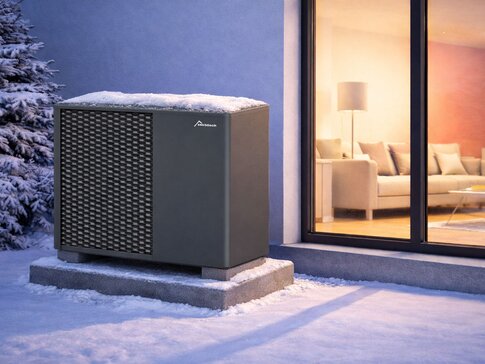 Monoblock-Luft-Wasser-Wärmepumpe von bösch im Winter, mit Schnee bedeckt, auf Betonsockel neben Fensterfront eines modernen Einfamilienhauses