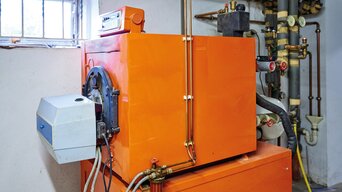 Ein orangefarbener Heizkessel in einem Kellerraum, flankiert von verschiedenen Rohrleitungen und Ventilen. Ein kleinerer, weißer Gerätekasten ist auf der linken Seite an den Kessel angeschlossen, und oberhalb des Kessels befindet sich ein alter Stromzähler. Im Hintergrund sind isolierte Rohre an der Decke zu sehen.